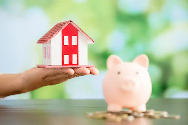 Mano sostiene una casa en miniatura junto a hucha cerdito, aludiendo a liquidación de bienes gananciales tras divorcio.