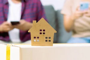 Figura de casa sobre caja de mudanza con pareja al fondo, simbolizando la vivienda familiar en liquidación de gananciales.