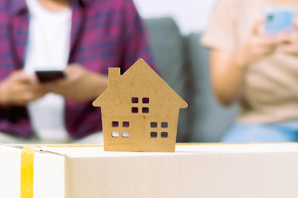 Figura de casa sobre caja de mudanza con pareja al fondo, simbolizando la vivienda familiar en liquidación de gananciales.